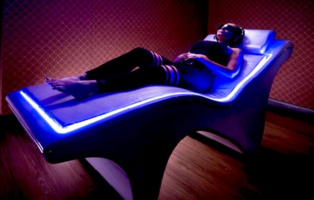 Person relaxing on a VEMI Bed with headphones and an eye mask, lit by the bed's blue edge lighting